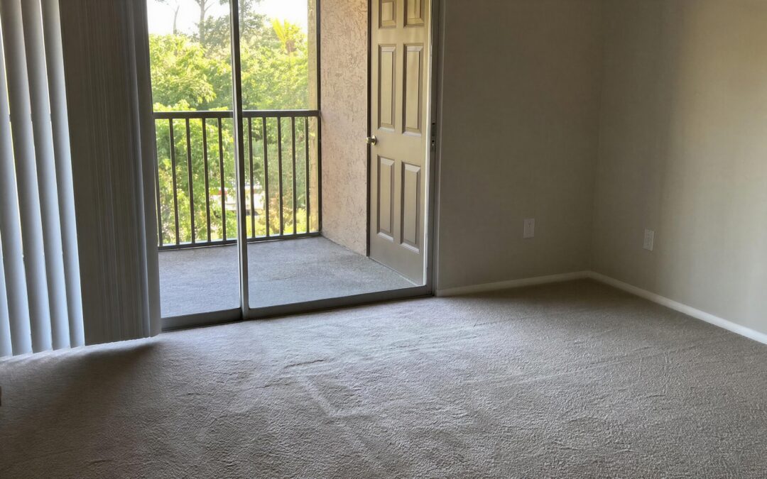 Empty apartment interior with beige carpet, sliding glass door open to a screened balcony with a view of trees outside, off-white walls, no furniture.